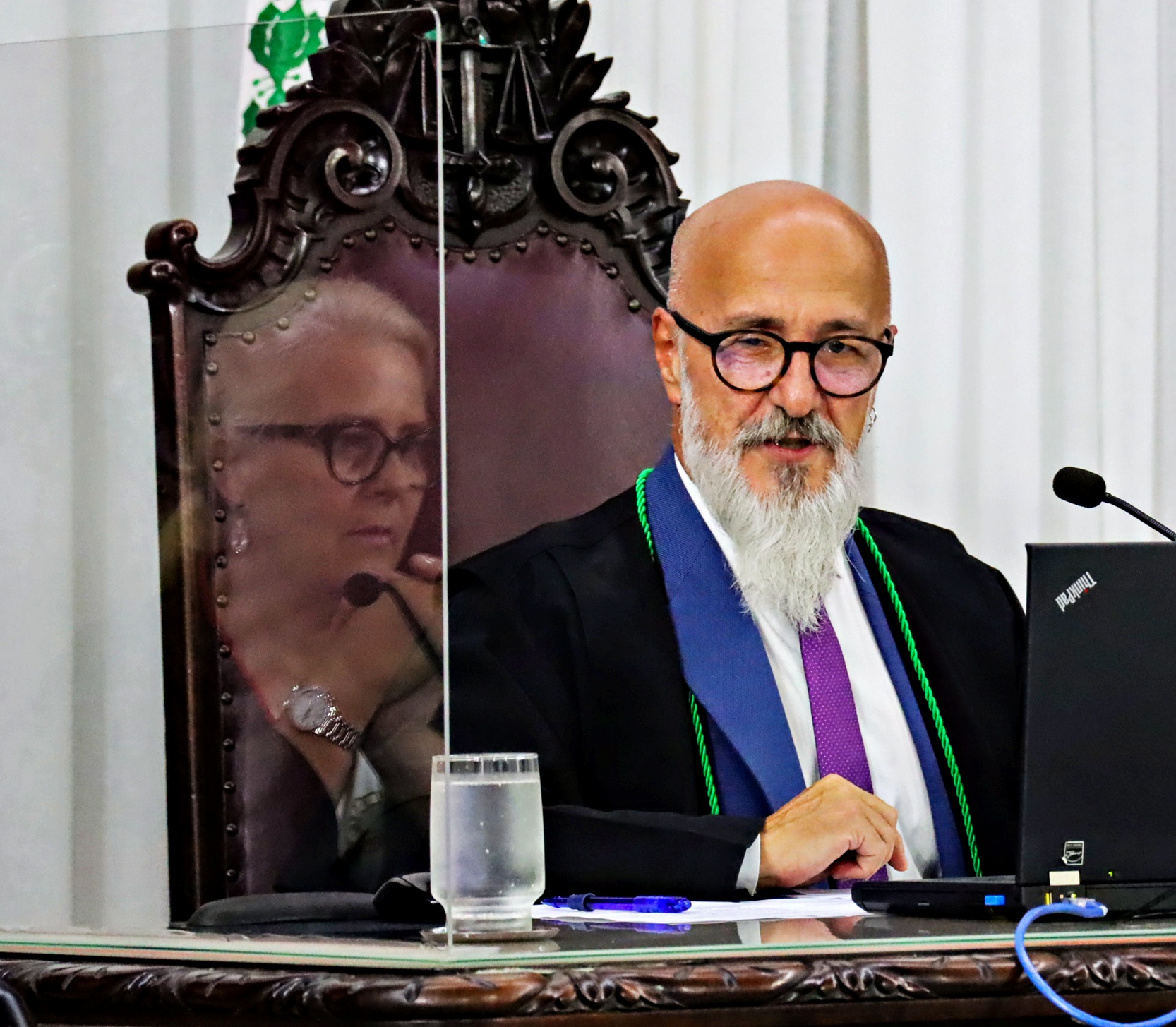 O conselheiro Fernando Guimarães comanda sessão presencial do Tribunal Pleno do TCE-PR; na imagem refletida na divisória, a procuradora-geral do Ministério Público de Contas (MPC-PR) Valéria Borba, que senta ao seu lado. Foto: Fabiano Contador/TCE-PR